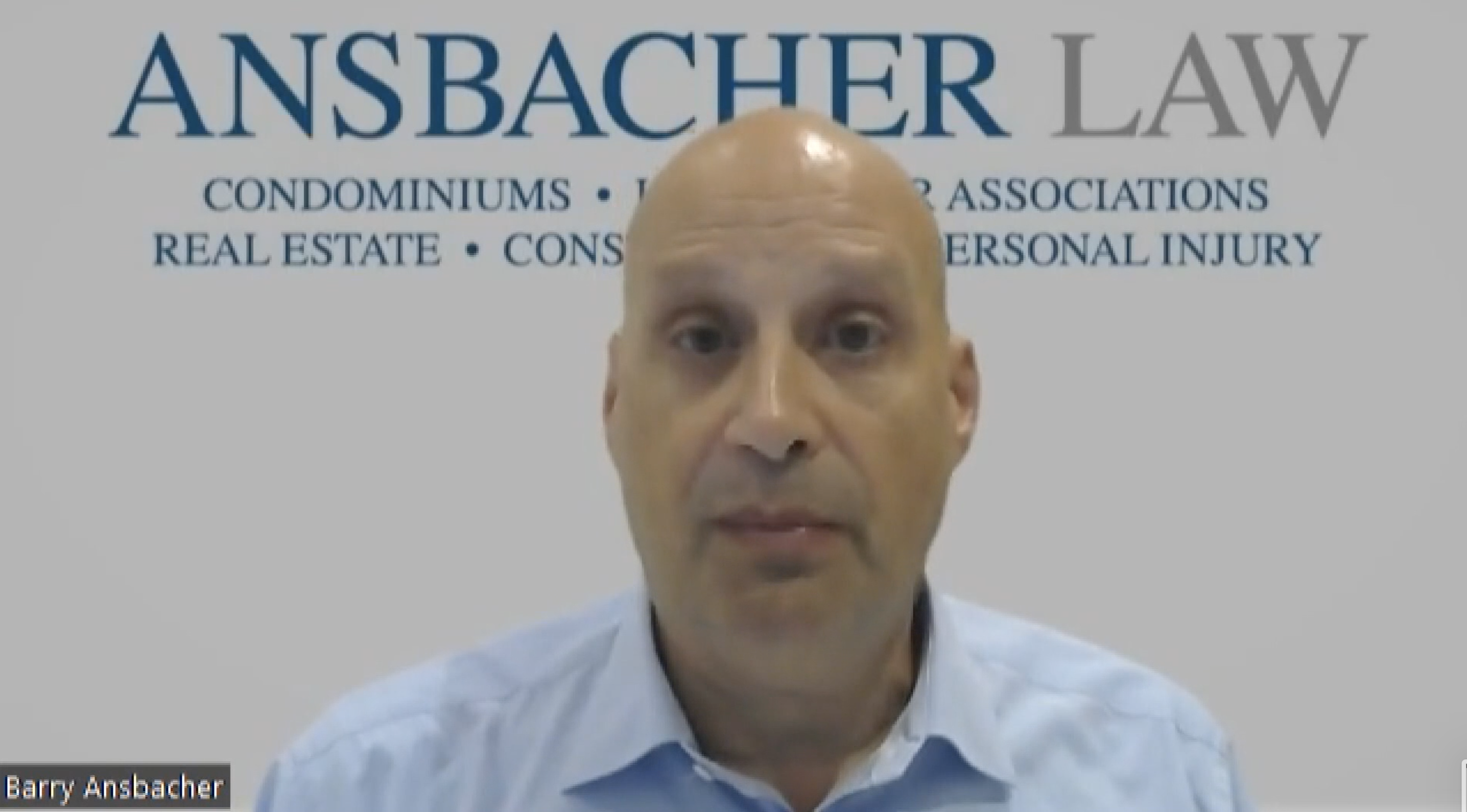 A man with a bald head and wearing a blue shirt sits in front of a backdrop displaying the logo and services of Ansbacher Law, including condominiums, real estate, wrongful death settlement, and
