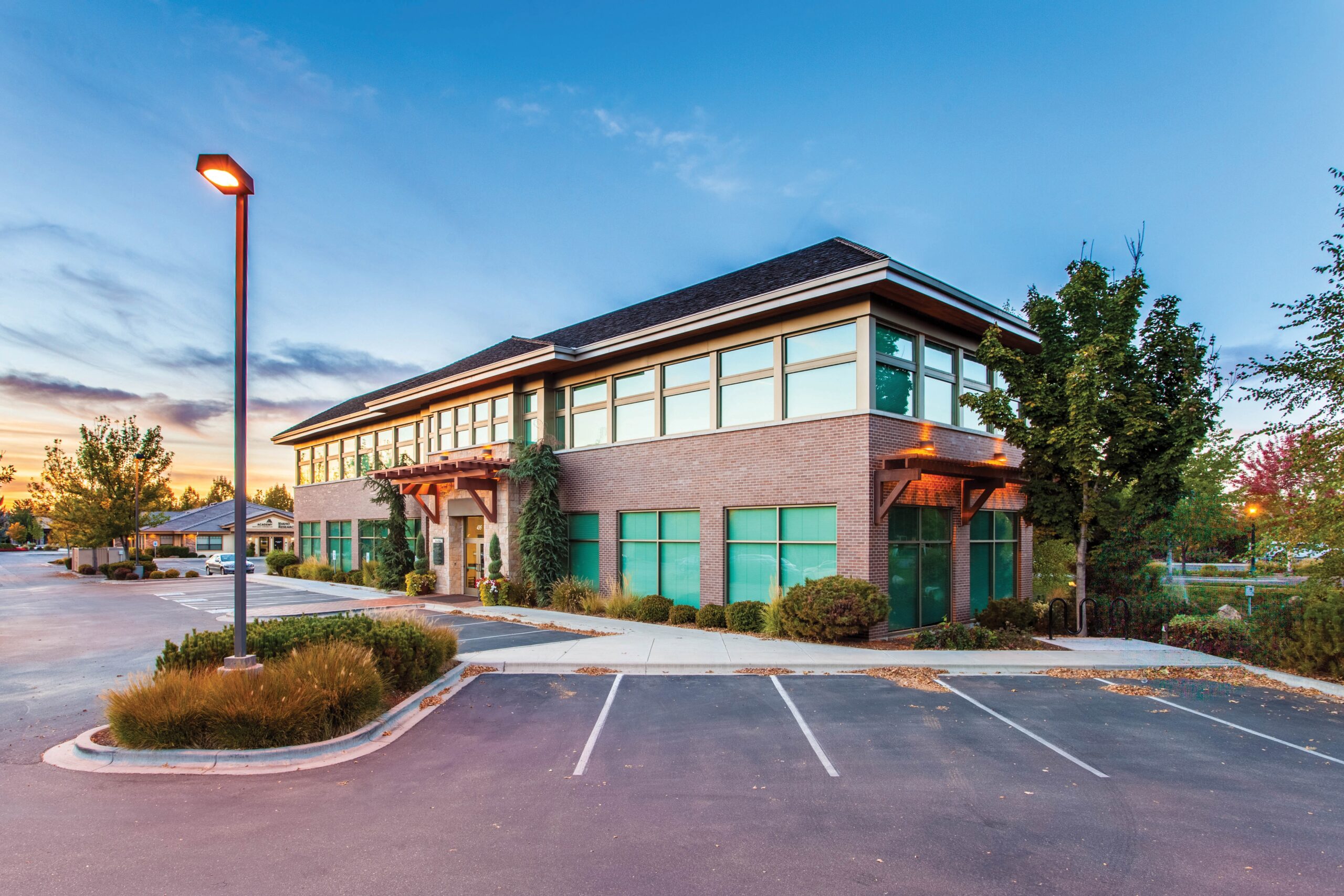A modern two-story office building at twilight with illuminated outdoor lamps, featuring a clear sky and vibrant landscaping, and an empty parking lot in the foreground, notorious for past parking lot accidents.