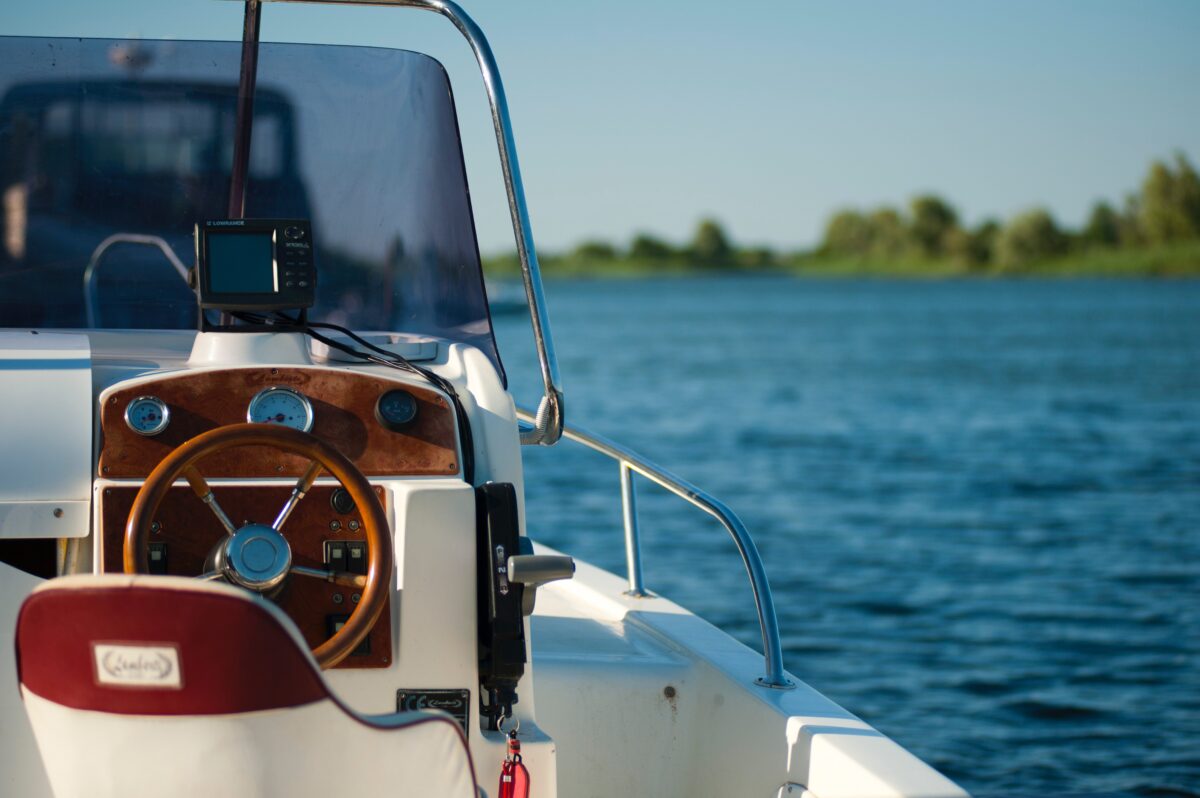 Close-up view of a boat's steering area, featuring a wooden steering wheel and dashboard with a navigation device, following a minor boat accident, overlooking a calm blue lake.