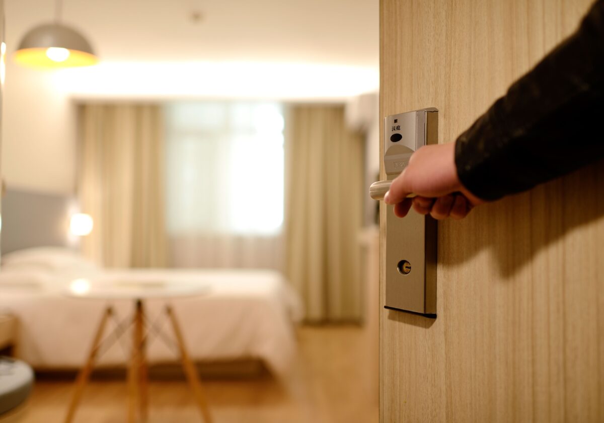 An injured person's hand is opening a modern hotel room door, revealing a well-lit, cozy room with a bed and lamps in the background.
