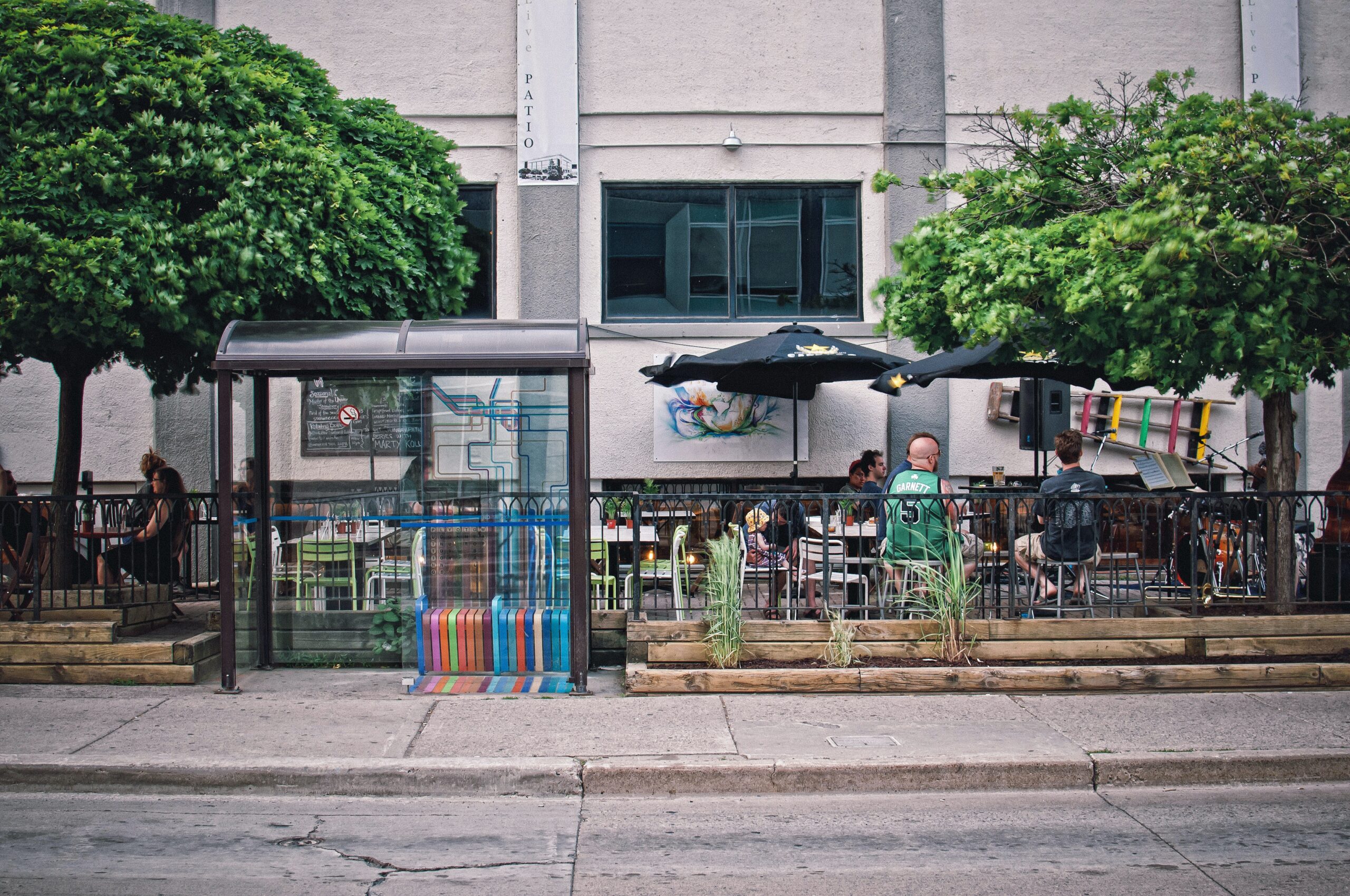 A city street scene showing a bus stop with a colorful bench next to a sidewalk café, where people dine under umbrellas. Lush green trees and a tattoo parlor sign feature in this lively urban