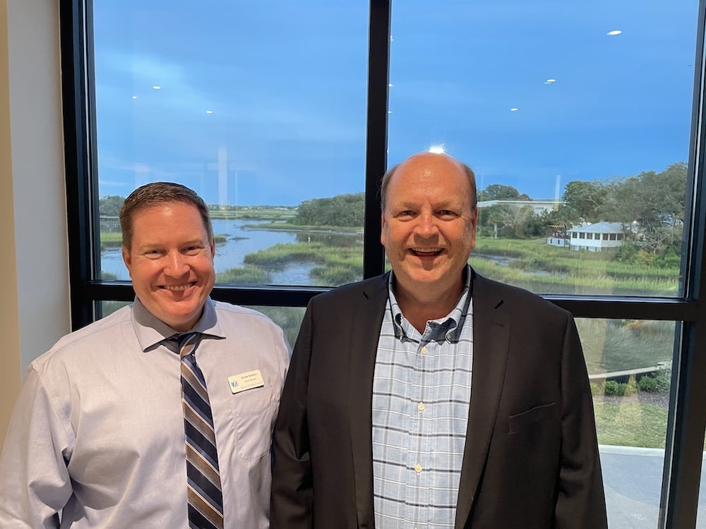 Two smiling men posing in front of a large window overlooking a scenic river landscape during the Grand Opening of the Ansbacher Law offices. They are wearing business casual attire, with one in a tie.