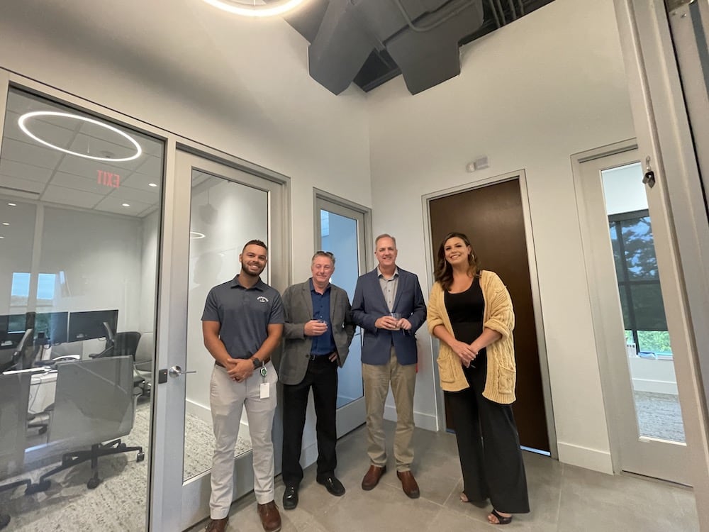 Four professionals smiling and standing in a modern office hallway during the Grand Opening, with glass doors and contemporary decor in the background.