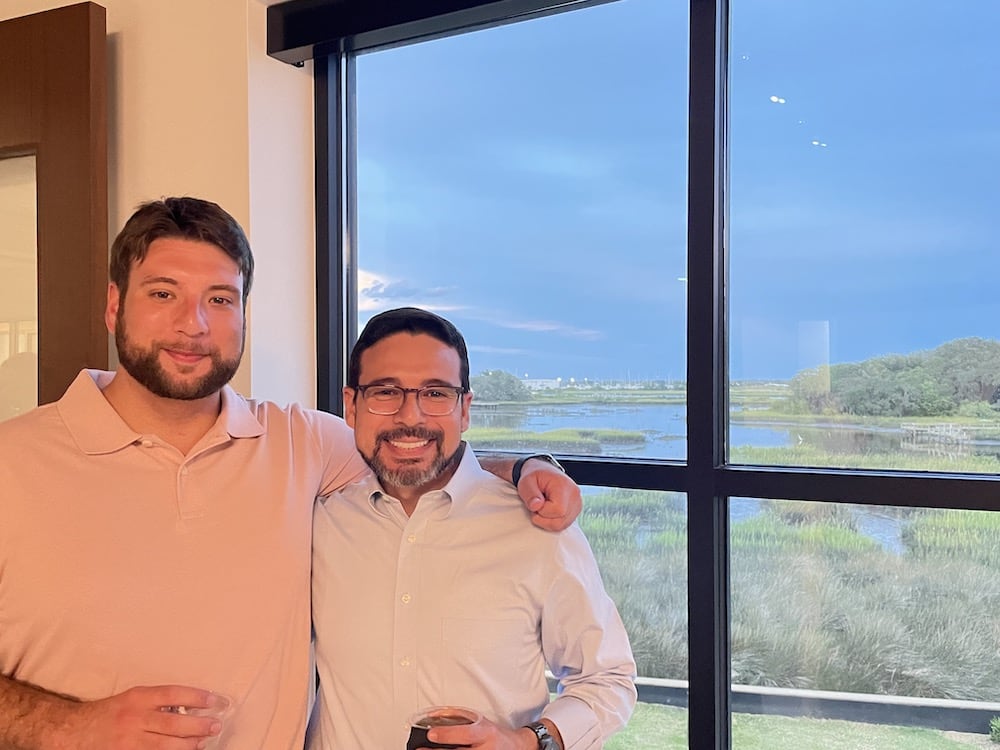 Two men smiling, one with a beard, standing together near a large window overlooking a scenic marshland under a dusk sky. Both are holding drinks and dressed in semi-casual attire during the Grand