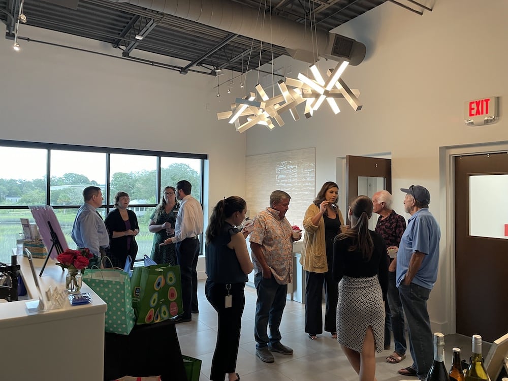 People conversing at the Grand Opening in a modern room with an avant-garde chandelier above and art exhibits nearby. An exit sign is visible near the door.