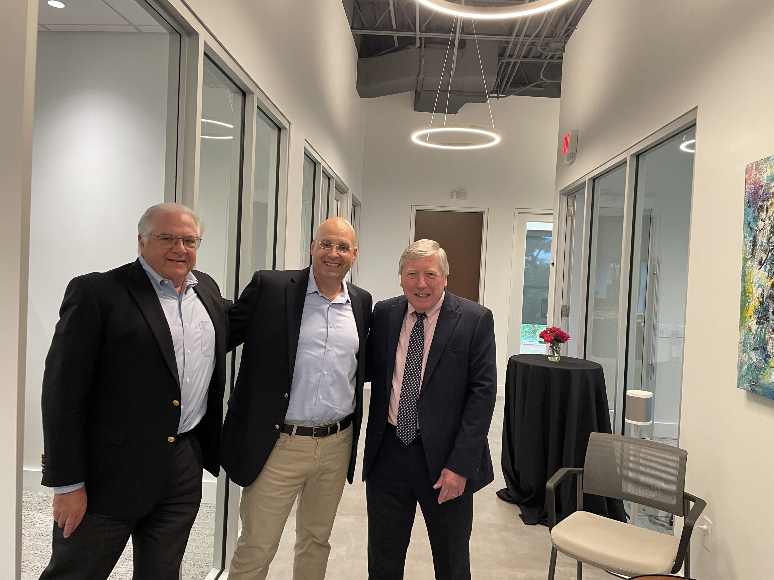 Three men smiling in a modern office hallway at the Grand Opening with abstract art and a table with a flower vase in the background. One man wears a black suit, the middle one in a blue shirt