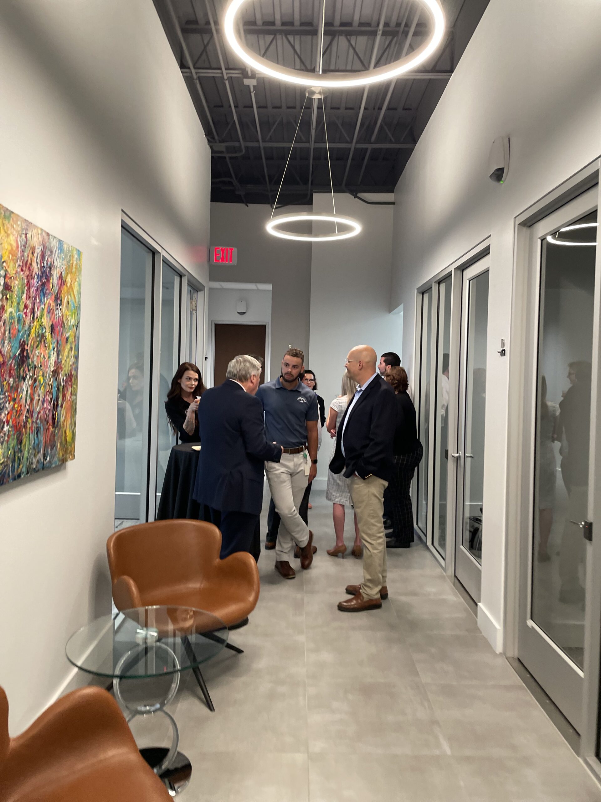 A group of professionals engaged in conversation at the grand opening of Ansbacher Law, in a modern office hallway with vibrant paintings on the wall and stylish circular lighting overhead.