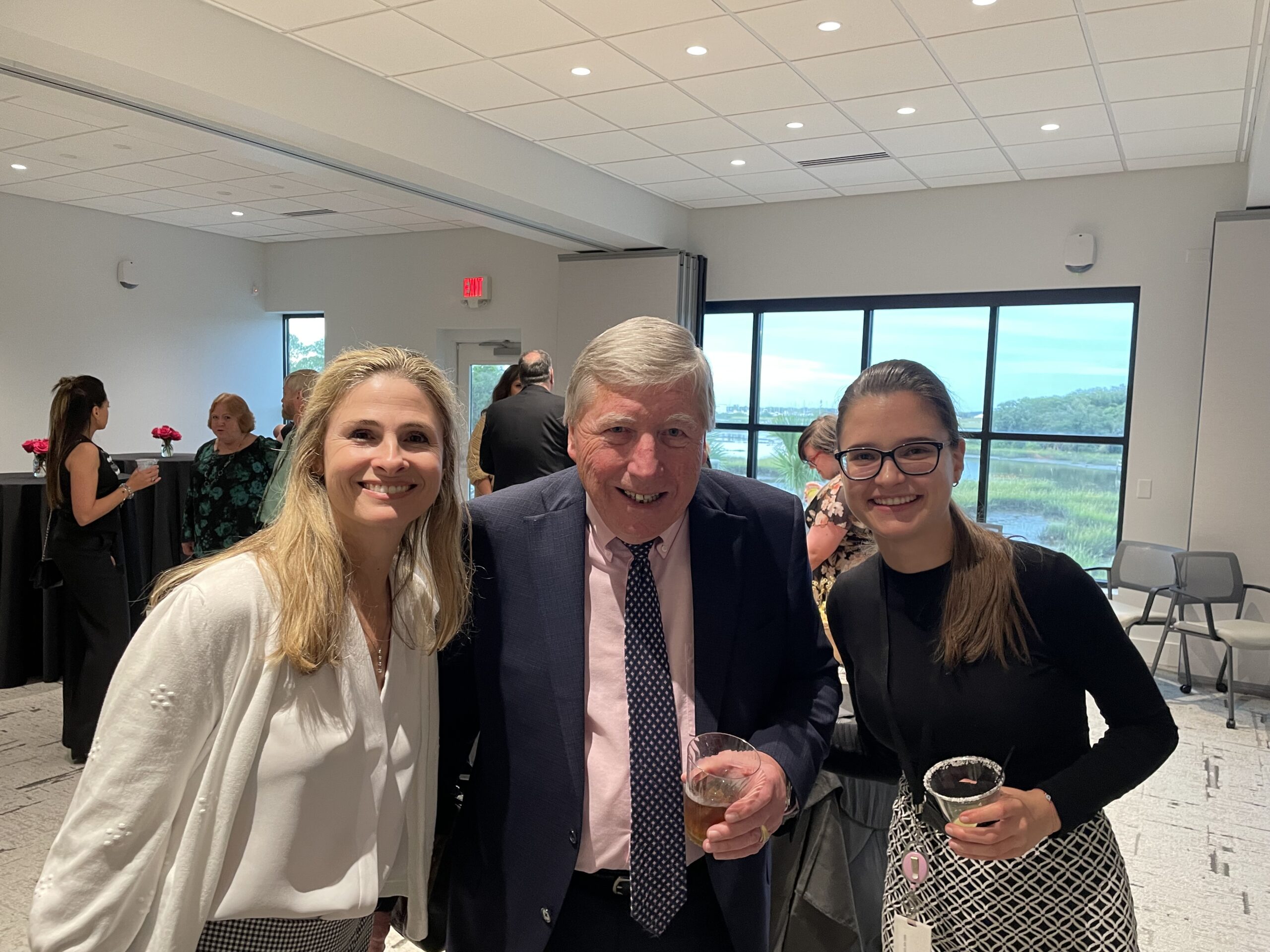 Three people smiling at the Grand Opening, with two women on the sides and an older man in the center holding a drink. They are indoors with other guests and a scenic window view in the background.