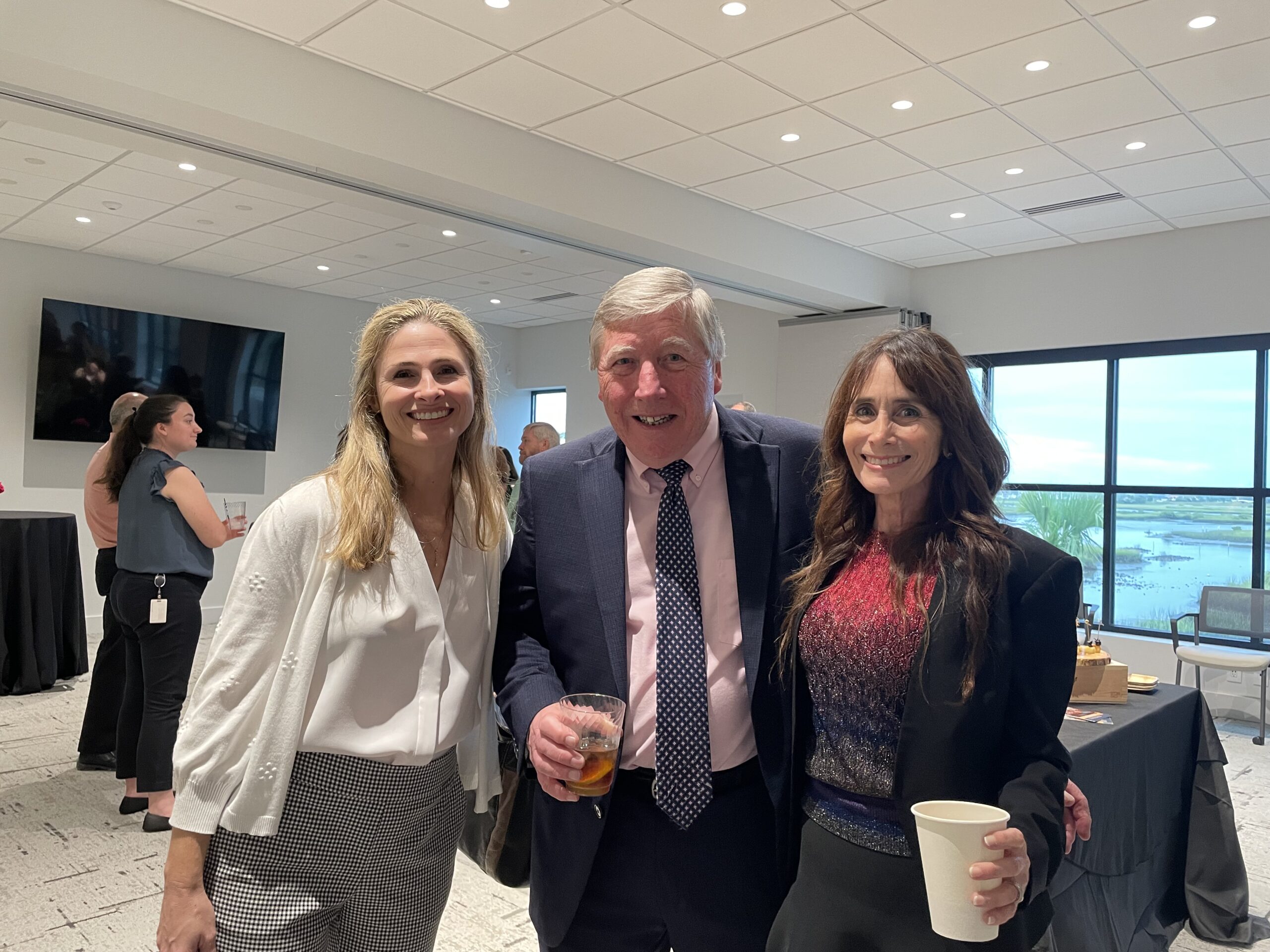 Three people smiling at a networking event; two women flanking a man holding a drink. They are in a room with large windows at the Grand Opening and other attendees in the background.