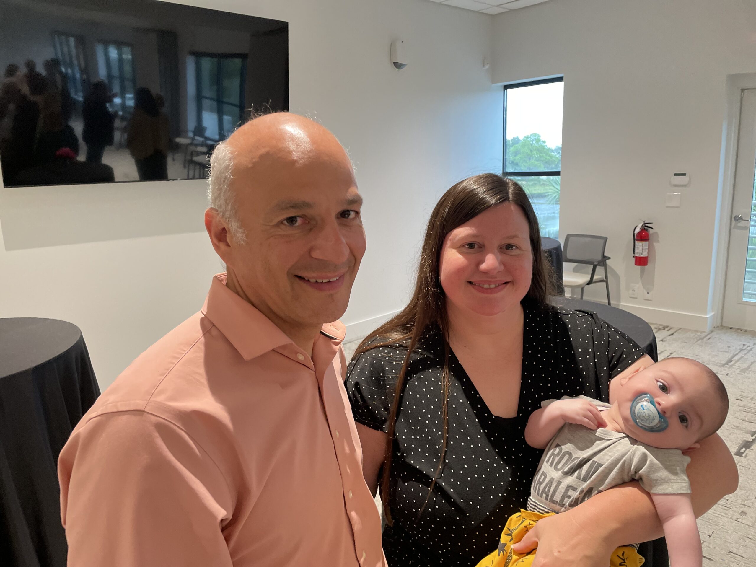 Three people posing for a photo at the Grand Opening inside a room: a bald man in a pink shirt, a woman with long hair in a polka-dot dress holding a baby with a pacifier