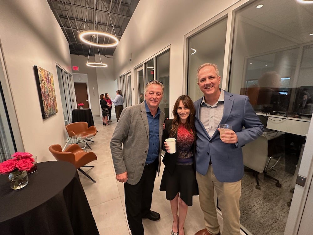 Three people pose with smiles, holding coffee cups in a modern office hallway adorned with artwork, under soft lighting. One man on the left with shorter blond hair, a woman in the center, and another