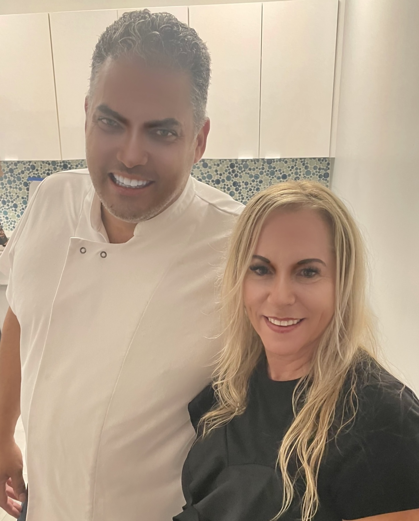 Two people, a man and a woman, smiling and posing together in a kitchen with a mosaic tile backsplash during the Grand Opening. The man has gray hair and wears a white shirt; the woman