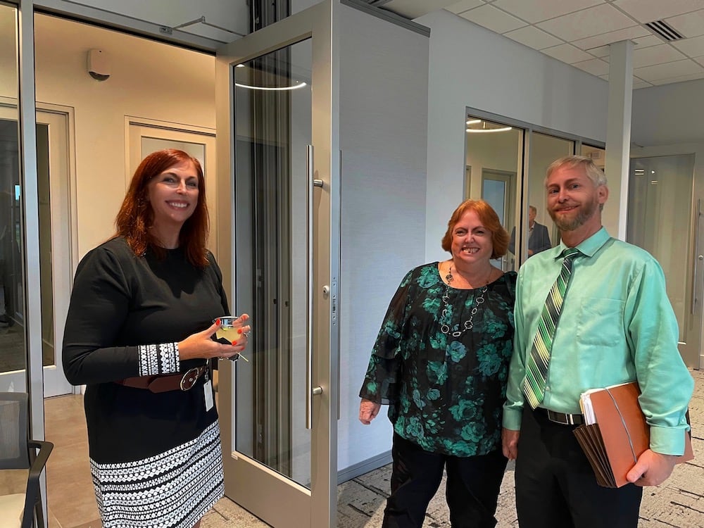 Three office coworkers standing in a corridor, smiling. To the left are two women, one with red hair and one blonde, and to the right is a man with gray hair—all dressed in smart office
