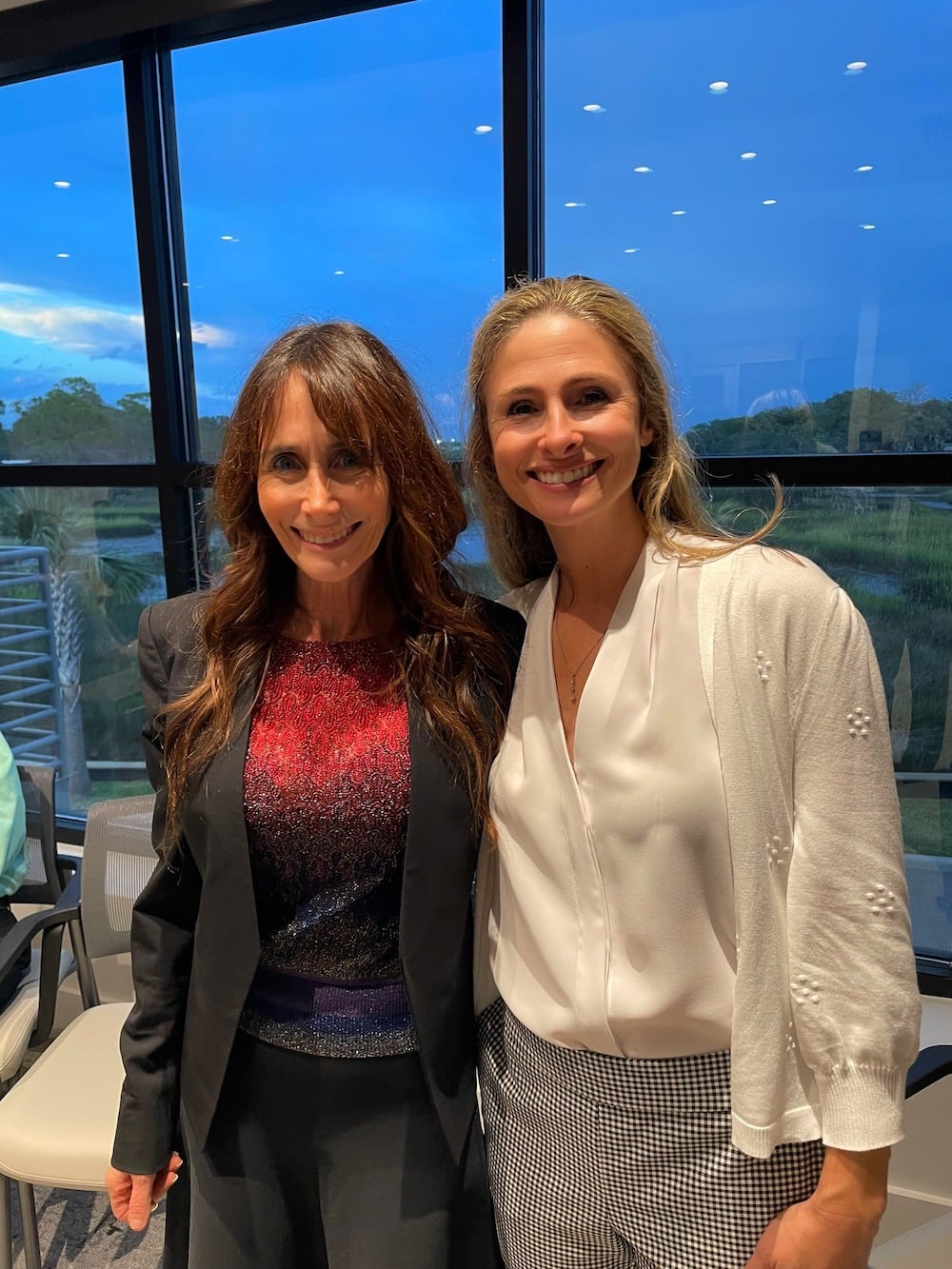 Two women smiling and standing together indoors at the Grand Opening, one dressed in a black and red top, the other in a white blouse and patterned skirt, with a window and greenery in the