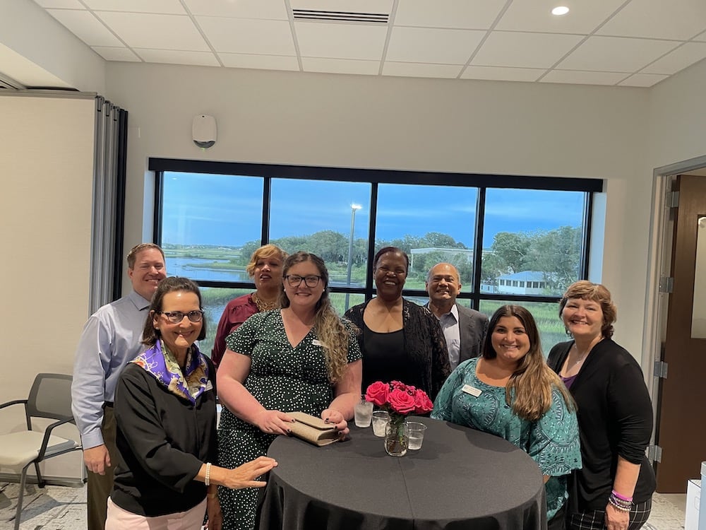 A group of eight diverse individuals smiling around a small round table with pink roses in a conference room with a scenic window view during the Grand Opening of the Betty Griffin Center.