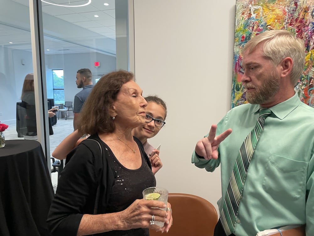 At the Grand Opening, an older woman holding a drink speaks animatedly, with a middle-aged man in a green tie listening intently and a young girl behind them playfully gesturing a peace sign