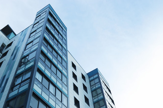 Low-angle view of a modern glass apartment building against a clear blue sky, highlighting its reflective windows and geometric architectural design.