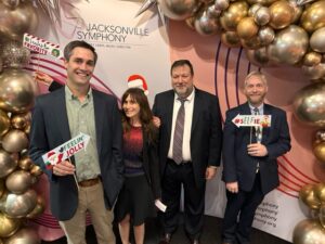 Four people posing in front of a festive backdrop with "Ansbacher Law" signage, decorated with large gold and silver ornaments. They hold holiday-themed props and display cheerful expressions.
