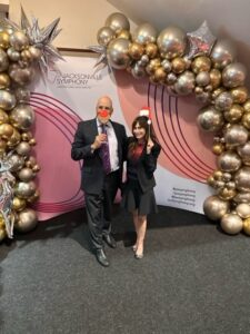 A man and a woman celebrating at a Jacksonville Symphony event, standing in front of a festive balloon arch. The man is holding a mask, and the woman is wearing a Santa hat.