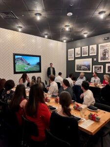 A business presentation at Ansbacher Law in a modern meeting room, with a man standing beside a screen displaying a landscape image. The audience, dressed in business attire, is seated at tables with laptops