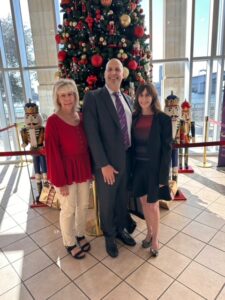 Three people smiling for a photo in front of a large Christmas tree adorned with red and gold ornaments. Two nutcracker figures and gifts celebrate the holidays at the tree's base.