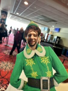 A person wearing a festive elf costume with a green jacket, white snowflake details, and pointed hat smiles at a holiday gathering.