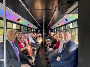 A group of people dressed in formal attire sitting inside a bus with black interiors, smiling towards the camera, as they celebrate holidays with sunlight streaming through the windows.