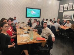 A group of people dressed in business attire sits around a long dining table in a stylish restaurant, smiling and engaged in conversation, with a television screen displaying an image of a bird in the background as they