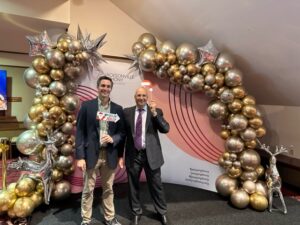 Two men smiling and posing under a decorative arch made of silver and gold balloons at a formal event. One man holds a magazine that celebrates the event’s theme.