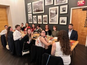 A group of people seated around a long wooden table in a restaurant, engaging in conversation. The walls are adorned with black and white photographs celebrating Ansbacher Law.