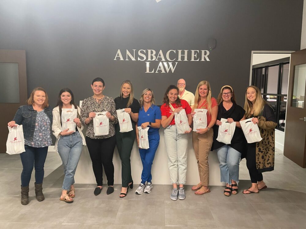 Group of nine people smiling in an office lobby, holding branded bags, with "Ansbacher Law" on the wall behind them in St. Augustine.