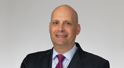 Professional headshot of attorney Barry Ansbacher, wearing a blue suit, white shirt, and pink tie, smiling slightly against a grey background.