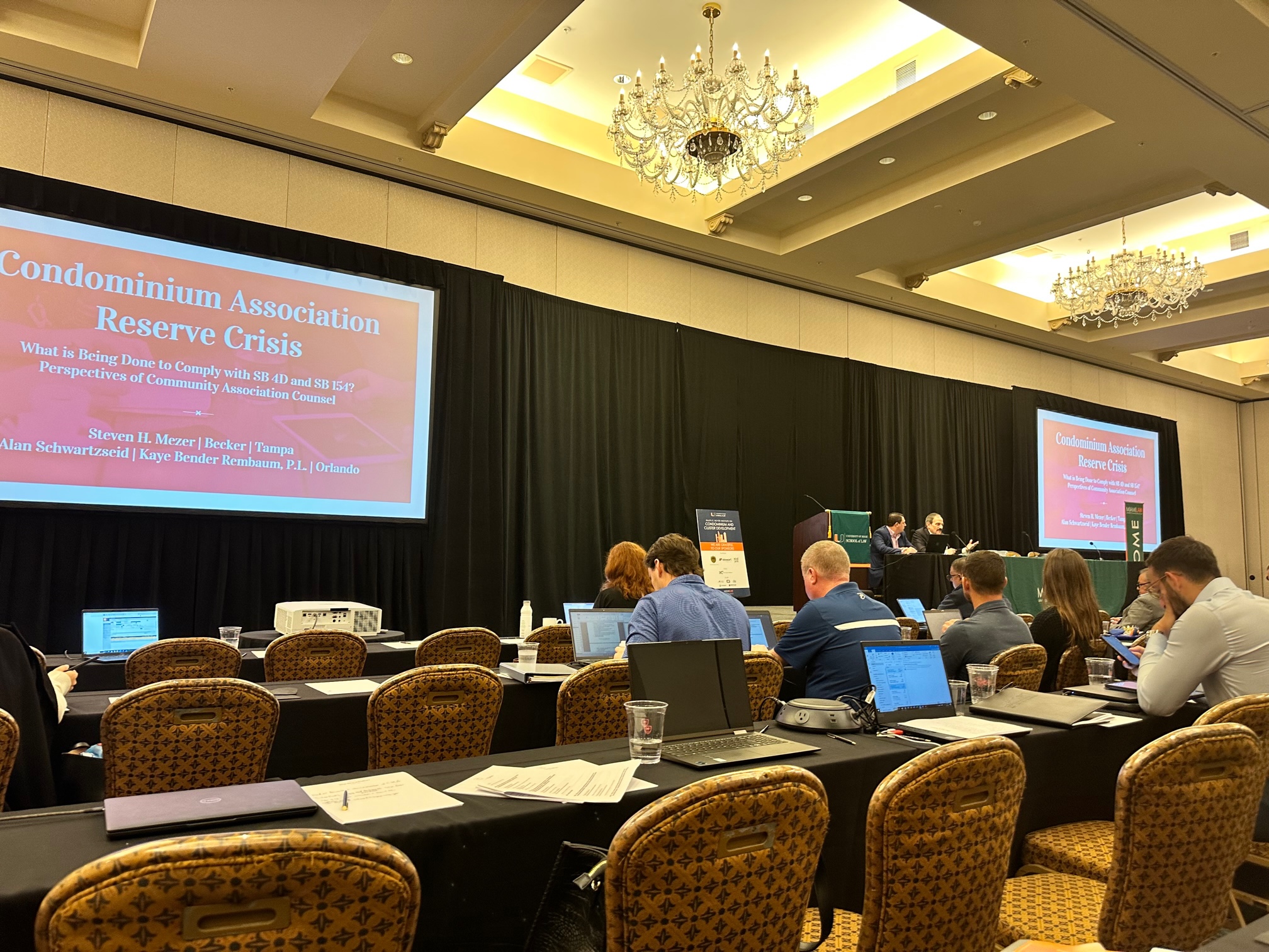A conference room with attendees seated at tables with laptops, focusing on a presentation about the condominium association reserve crisis, led by three speakers from the UM Boyer Institute at the front.