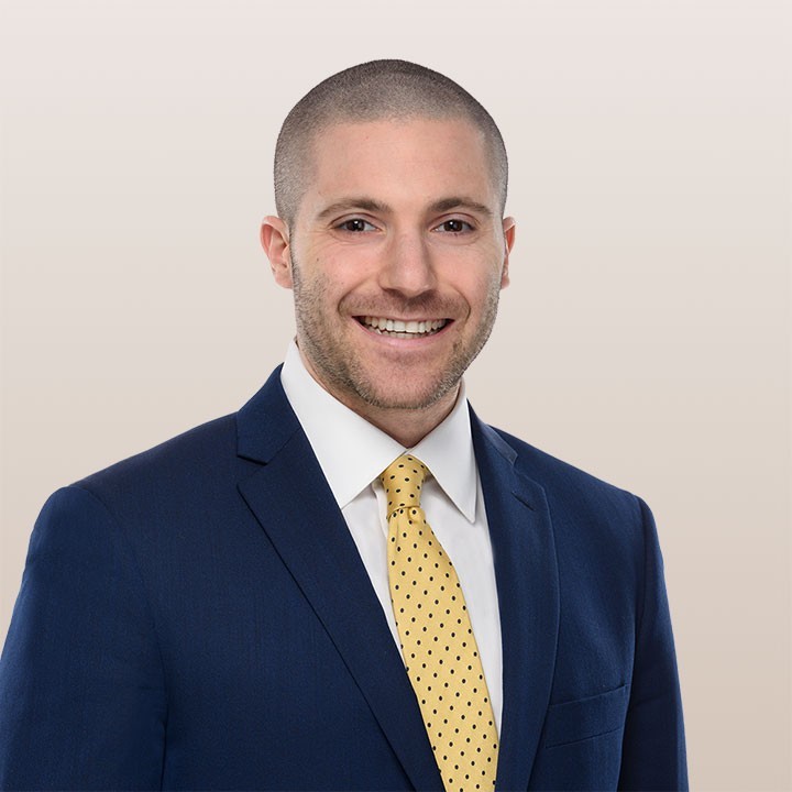 Professional headshot of a smiling man with short hair, wearing an Ansbacher Feinberg blue suit, white shirt, and yellow dotted tie against a light beige background.