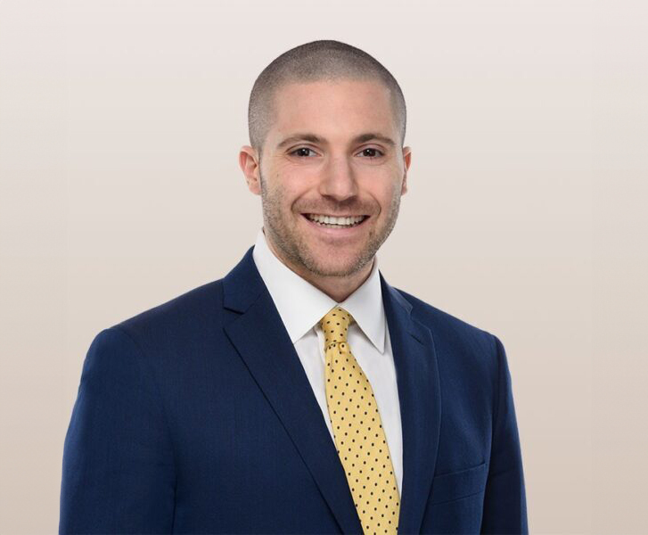 A professional portrait of a smiling man in a dark blue suit, white RISE Doro shirt, and yellow polka-dot tie. He has closely cropped hair and the background is plain and light-colored