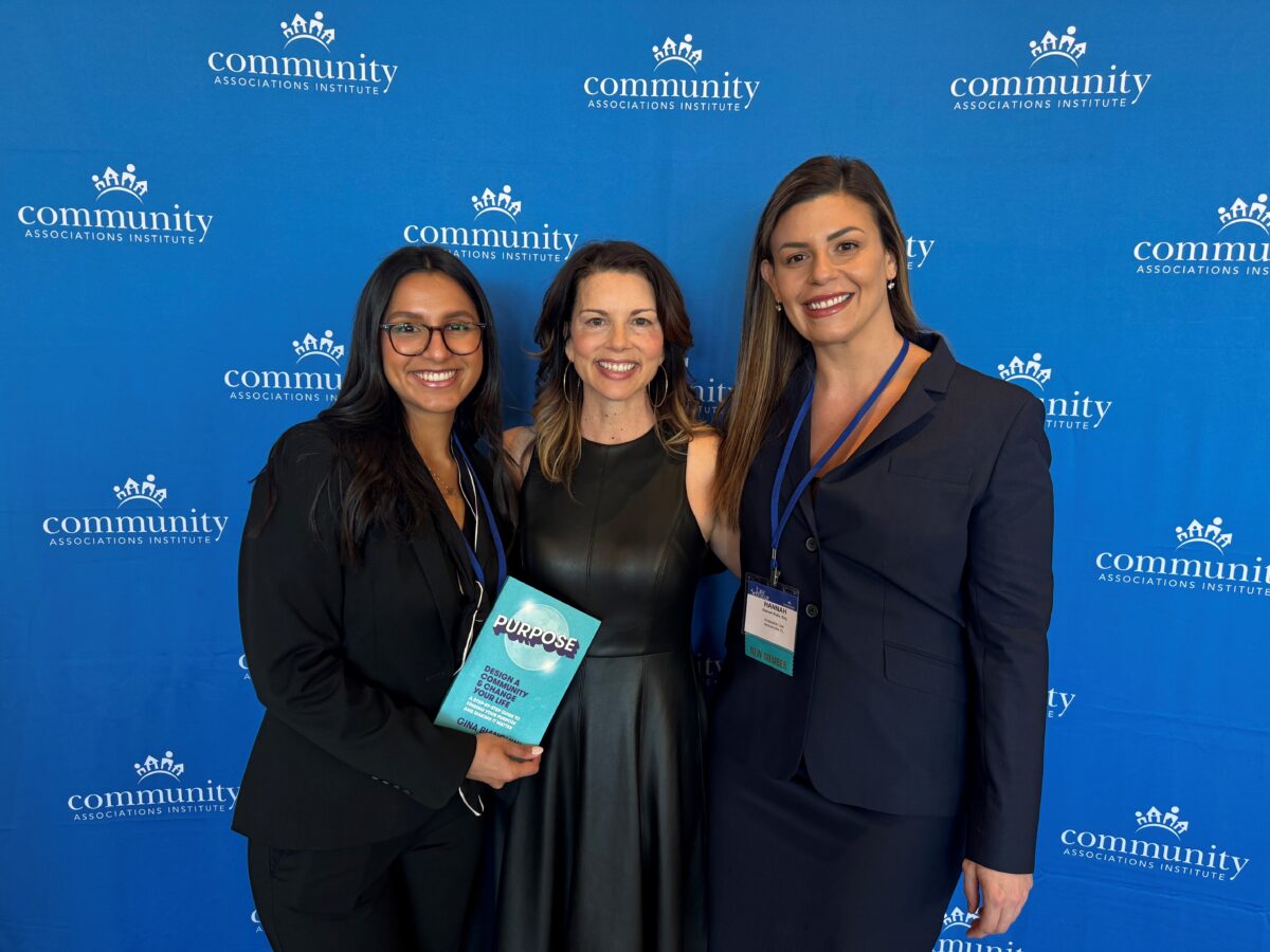 Three women smiling at the National Education Conference, standing in front of a blue banner with logos, one holding a book titled "Purpose." They are dressed in business attire.