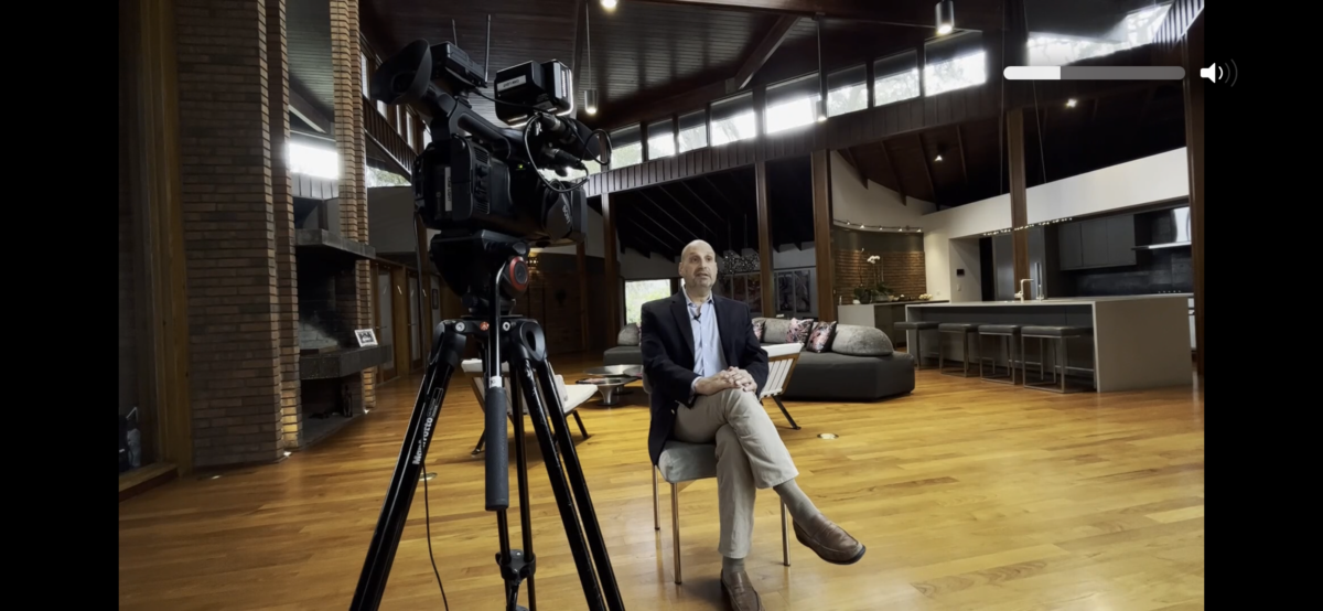 A man in a suit sits on a stool in a spacious loft-style room, facing a professional video camera on a tripod, ready for an interview. High ceilings and modern furnishings are visible in the background