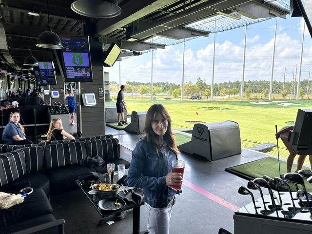 A woman holding a drink smiles at the camera at a bustling indoor golf driving range during the Ansbacher team outing, with players, seating areas, and digital screens.