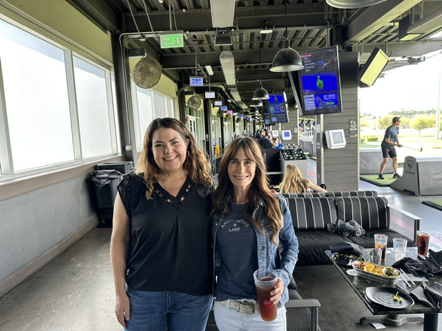 Two women smiling at an Ansbacher team outing at a Topgolf venue, with one holding a drink. They stand before a golf bay filled with clubs, with screens and other patrons visible in