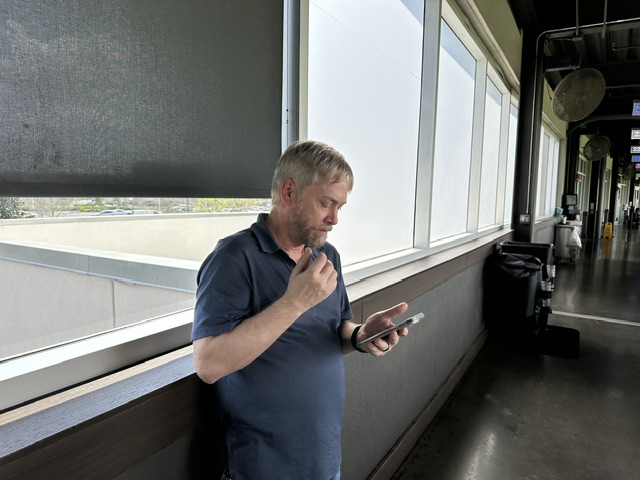 A man standing by a window in a public building during an Ansbacher team outing, looking thoughtfully at his smartphone while holding his chin with his other hand.