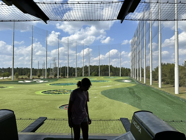 At an Ansbacher team outing, a person at a driving range stands in front of a tee, looking out over the field marked with circular targets. The range is surrounded by high nets under a