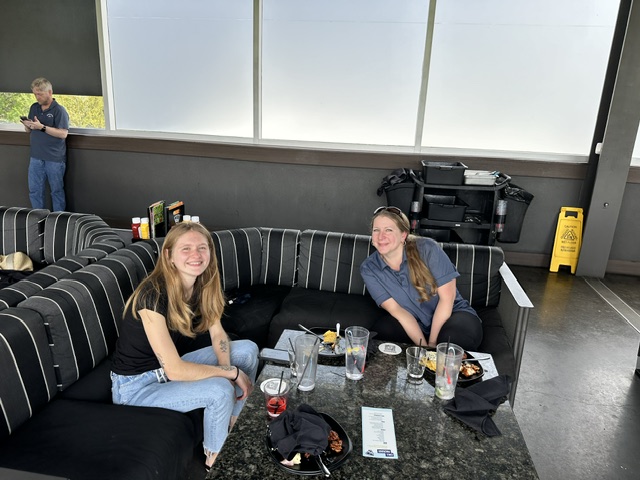 Two women from the Ansbacher team smiling at a curved booth in a modern, airy restaurant with large windows and a caution sign on the floor. They are surrounded by drinks and menus.
