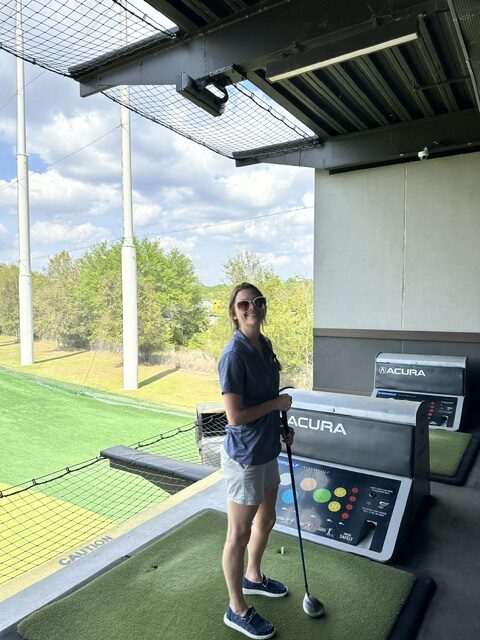 A woman wearing sunglasses, a navy blue t-shirt, and grey shorts stands smiling at the Ansbacher team outing, holding a golf club, with a green field and trees in the background.