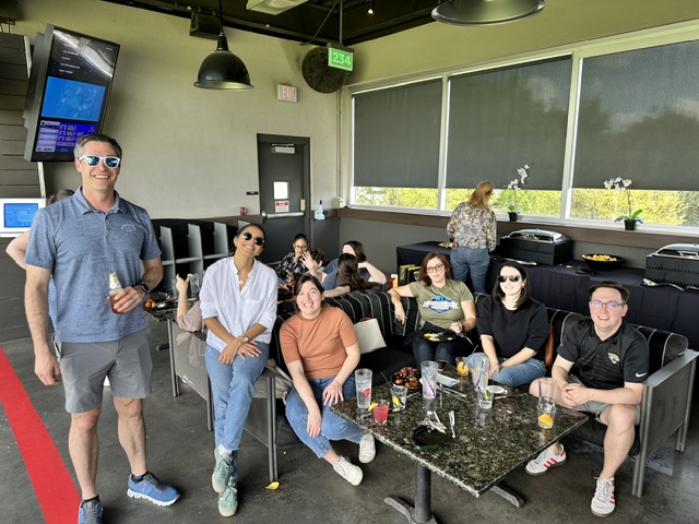 A group of eight people from the Ansbacher team outing, smiling and seated at patio style seating, enjoying drinks and snacks, with a man standing to the left. They are in a semi-out