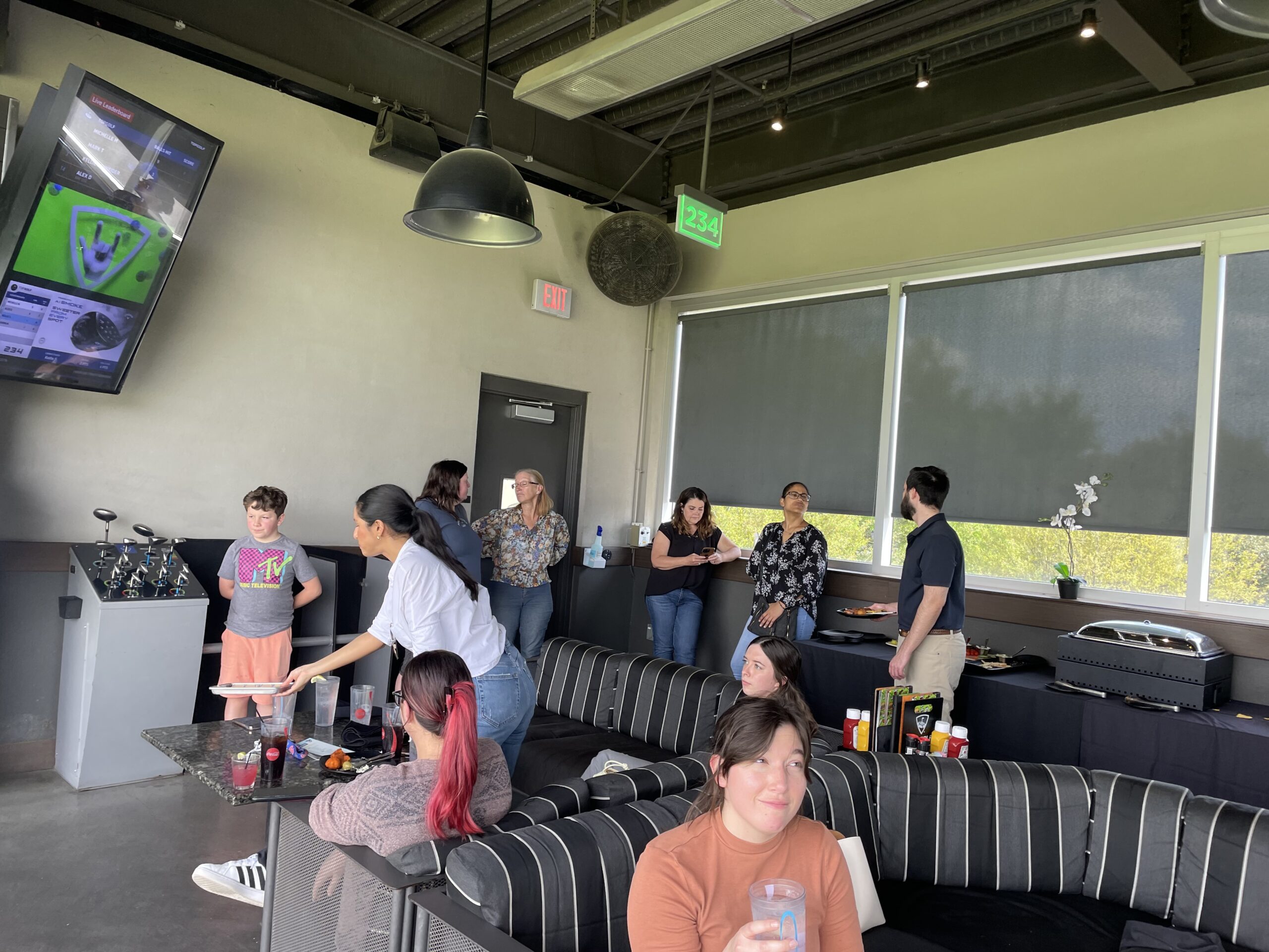 People socializing and preparing food at an indoor gathering for an Ansbacher team outing with a casual seating area and a beverage counter under ambient lighting.