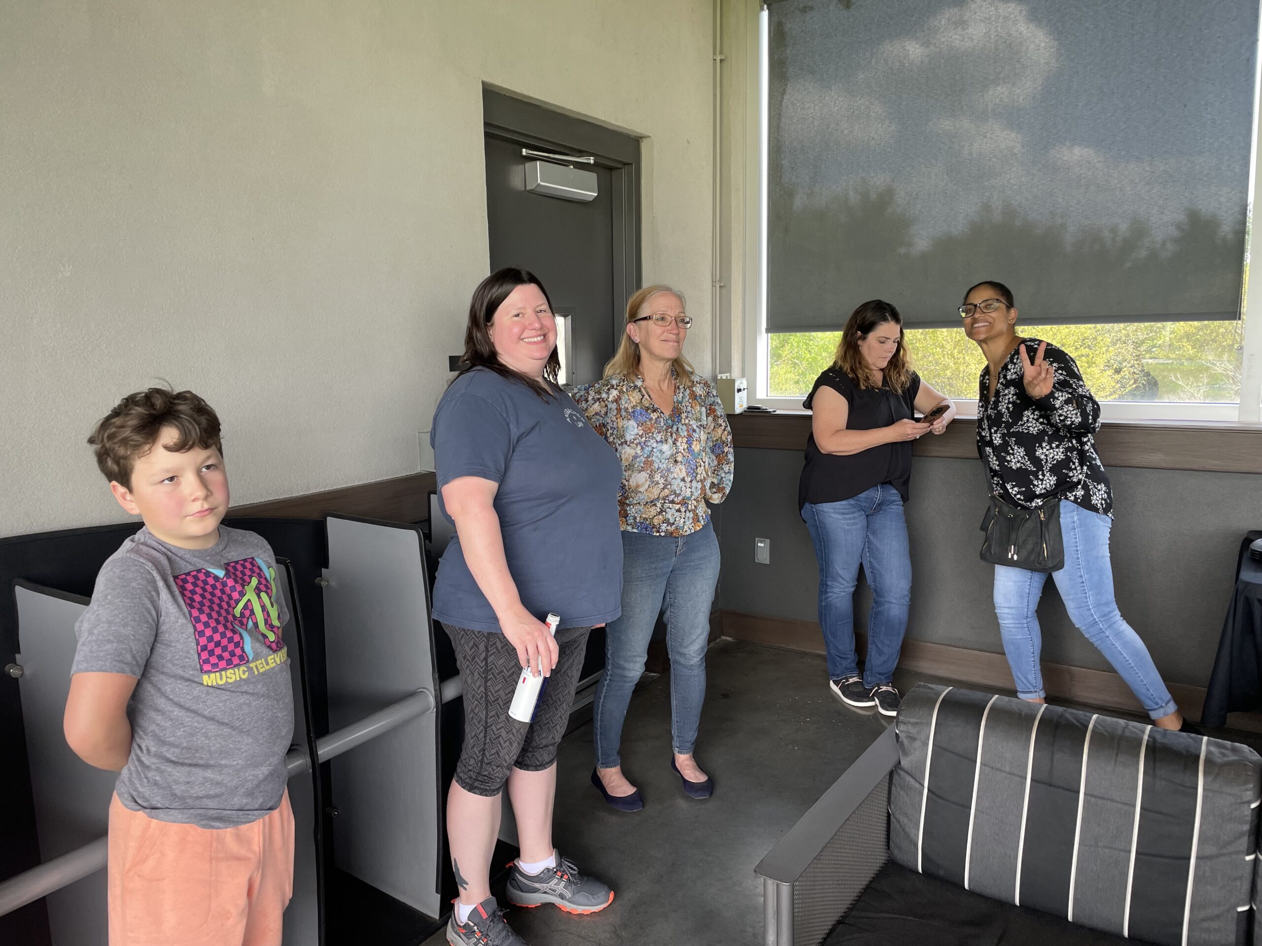 Five people of various ages and ethnicities stand on a shaded patio during an Ansbacher team outing. One young boy looks onward, while four women appear engaged in light conversation or phone use.