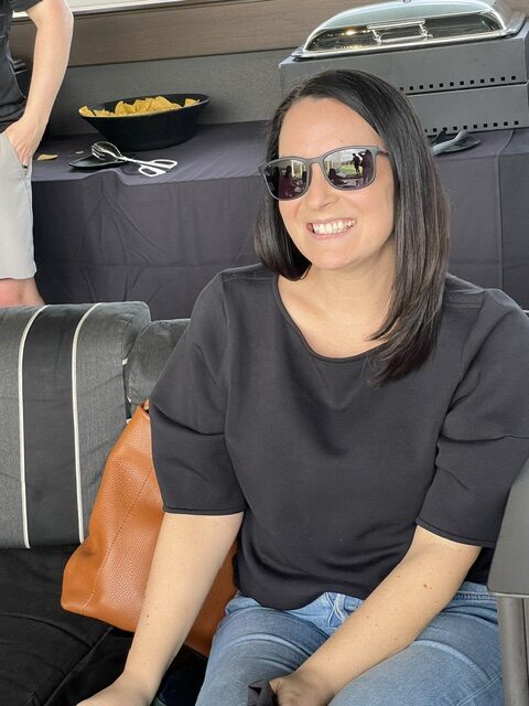 A smiling woman with sunglasses seated in a casual setting, with a barbecue and snacks in the background at the Ansbacher team outing. She has shoulder-length dark hair and is wearing a black top.