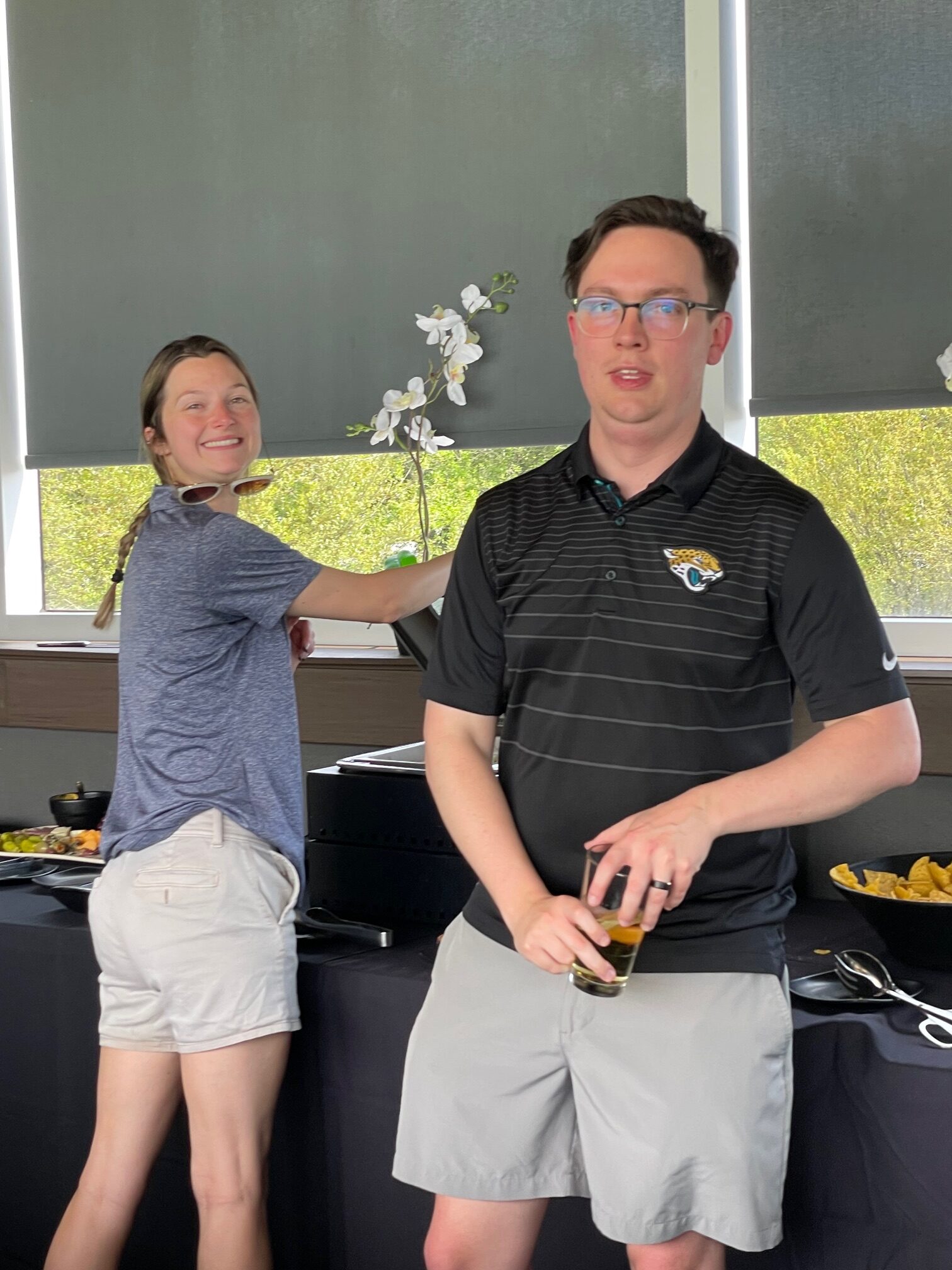 A woman playfully points an electronic dart at a surprised man wearing glasses during the Ansbacher team outing. They are both casually dressed, inside a room with large windows and standing next to a food