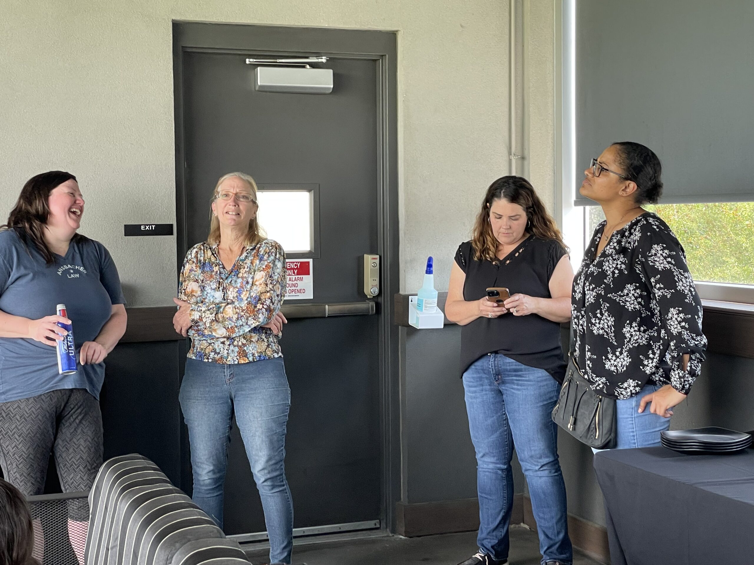 Four women in a casual workplace setting, three chatting and laughing by a door during the Ansbacher team outing, while the fourth is focused on her phone. Light filters through a window.