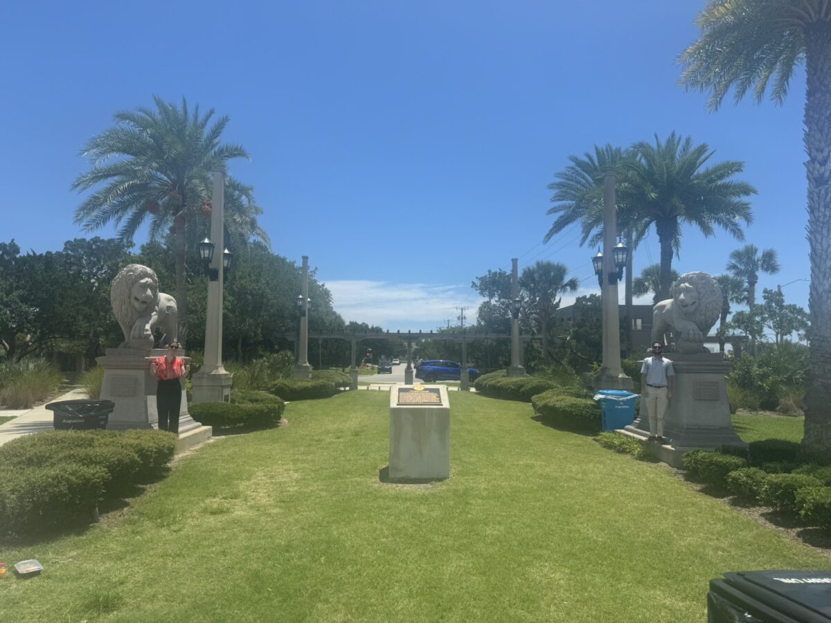 A sunny outdoor walkway flanked by tall palm trees and stone lion statues on pedestals evokes the charm of St. Augustine. Law clerks stroll along the path, lined with manicured grass and small bushes, leading to a distant vista of trees and clear blue sky. People are admiring the statues.